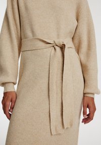 Robe en tricot côtelé beige avec une ceinture à nouer à la taille, manches longues et texture douce. Le tissu semble chaud et confortable.