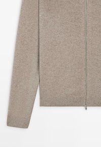 Hellbeige Strickjacke mit Reißverschluss, gefertigt aus einem weichen Strickstoff. Verfügt über einen gerippten Saum und Bündchen, eine glatte Textur und silberne Reißverschlussdetails.