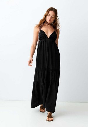 Mujer con vestido maxi negro sin mangas con tirantes de encaje, sandalias marrones con tachuelas y un collar de encaje a juego, de pie sobre un fondo blanco.