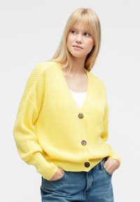 Κίτρινη πλεκτή ζακέτα με V-λαιμόκοψη, τρία καφέ κουμπιά, μακρύ μανίκι και άνετη εφαρμογή. Φορεμένη πάνω από λευκό τοπ και τζιν.