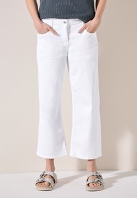 Pantalones anchos blancos con bolsillos delanteros, con cierre de botón y cremallera. Modelo con sandalias plateadas, de pie sobre un fondo neutro.