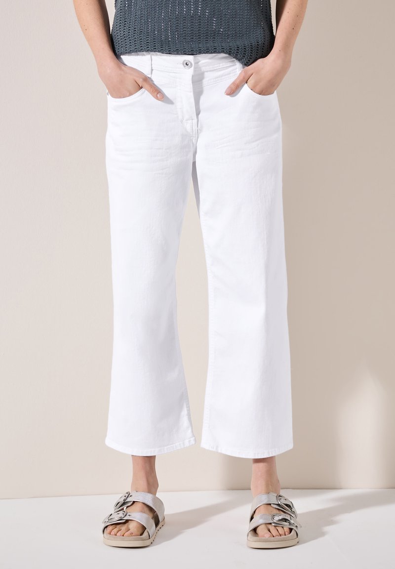 Pantalones anchos blancos con bolsillos delanteros, con cierre de botón y cremallera. Modelo con sandalias plateadas, de pie sobre un fondo neutro.