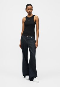 Γυναίκα που φοράει μαύρο αμάνικο τοπ, ευρύχωρο σκούρο τζιν και λευκά αθλητικά παπούτσια, στέκεται μπροστά σε απλό λευκό φόντο.