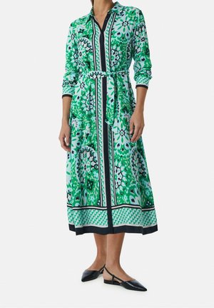 Femme portant une robe midi à manches longues, imprimé floral vert et blanc, avec une ceinture à la taille et des chaussures plates noires à bout pointu.