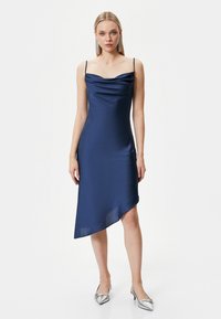 Marineblaues Satin-Kleid mit Cowl-Neck und dünnen Trägern, asymmetrischem Saum und glatter Textur, kombiniert mit silbernen spitzen Absatzschuhen.