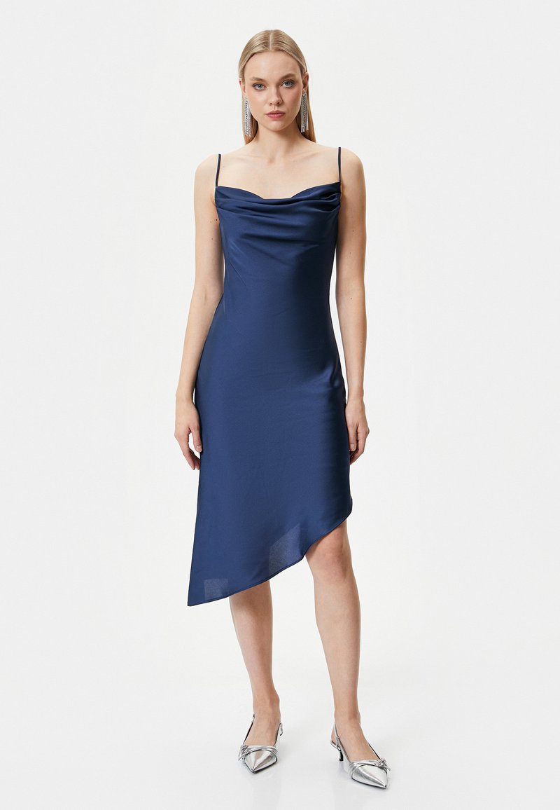 Marineblaues Satin-Kleid mit Cowl-Neck und dünnen Trägern, asymmetrischem Saum und glatter Textur, kombiniert mit silbernen spitzen Absatzschuhen.