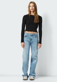 Juodas ilgomis rankovėmis ir banguotu apatiniu kraštu pasiekantis viršus, derinamas su šviesiai mėlynomis tiesiomis džinsomis ir smėliniais sportbačiais.