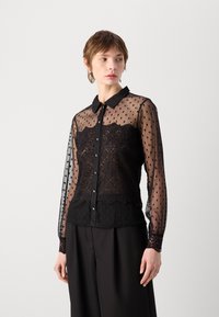 Chemisier noir transparent avec un motif à pois, détails en dentelle au niveau du corsage et manches longues. Il possède un col et une fermeture à boutons.