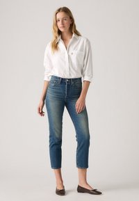 Balta marškinėliai su sagomis, aukšto liemens mėlyni džinsai su išbalinta apdaila, trumpas ilgis ir rudi baleto bateliai. Paprastas dizainas, laisvas stilius.