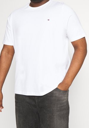 Uomo che indossa una t-shirt bianca a maniche corte senza motivi, con un piccolo logo rosso e nero sul petto, abbinata a jeans neri sbiaditi.