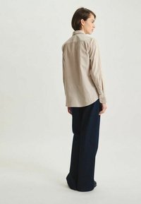 Chemise beige à boutons avec manches longues, détail de la pièce d'épaule arrière, associée à un pantalon en denim foncé à coupe ample. Tissu lisse, coupe ajustée.