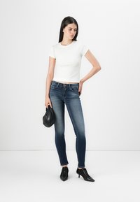 Fehér bordázott, rövid ujjú blúz, sötétkék skinny farmernadrággal párosítva. A modell egy fekete táskát tart és fekete, hegyes orrú magassarkút visel.