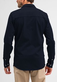 Man die een donkerblauw shirt met lange mouwen en beige broek draagt, vanaf de achterkant weergegeven tegen een effen achtergrond.