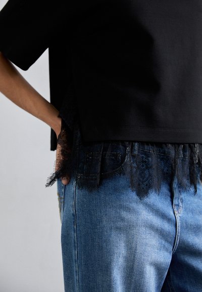 Czarny top z koronkowym wykończeniem na dole, noszony na niebieskich dżinsach, podkreślający kontrast faktur. Koronka ma kwiatowe wzory wzdłuż krawędzi.