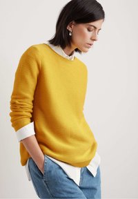 Gul ribbad tröja med rund halsringning, lagd över en vit skjorta med krage. Blå denimjeans med avspänd passform.