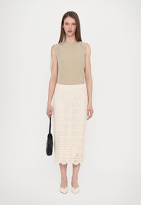 Top beige senza maniche abbinato a una gonna midi bianca in pizzo con motivi circolari, accompagnato da una borsa a tracolla nera intrecciata e tacchi beige appuntiti.