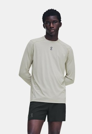 Homme à la peau foncée portant une chemise claire à manches longues et un short noir, debout les mains dans le dos sur un fond uni.
