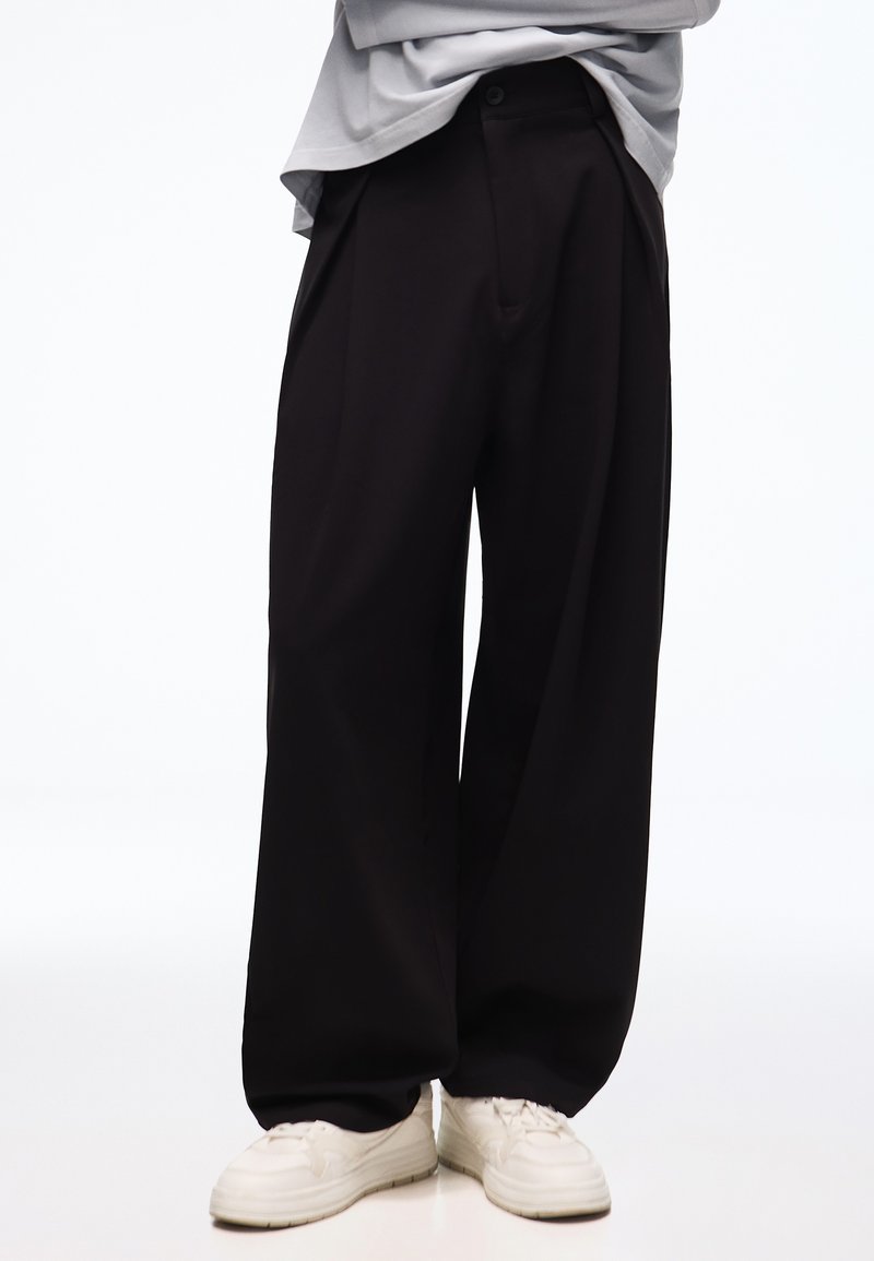Personne portant un pantalon noir ample, des baskets blanches et un haut gris, vue de la taille vers le bas sur un fond clair uni.