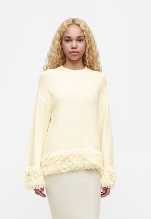 Woman with long curly blonde hair wearing a cream-colored sweater with textured fringe on cuffs and hem, standing against a plain background.
