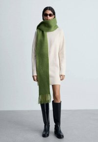 Sciarpa di lana verde con estremità frangiate, indossata sopra un vestito maglione beige chiaro. Stivali neri fino al ginocchio completano il look. Texture complessivamente liscia.