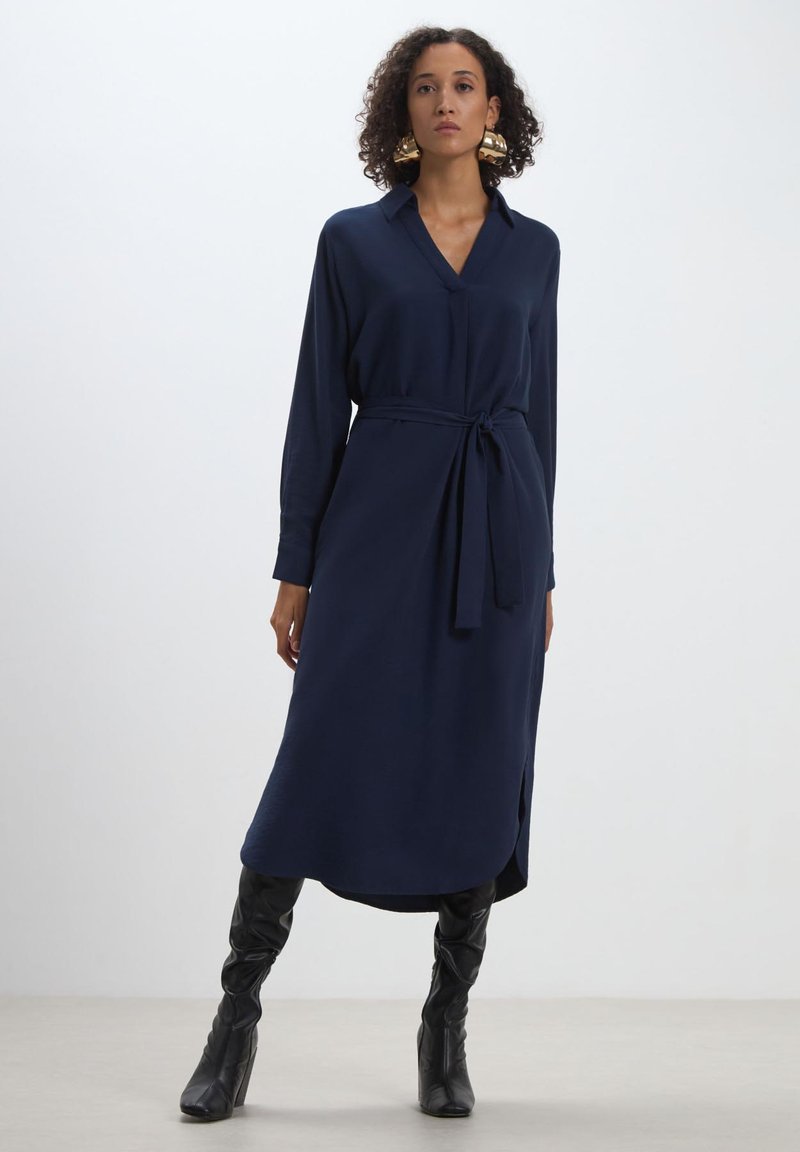 Robe en bleu marine à manches longues avec col, taille ceinturée et fentes latérales. Texture de tissu lisse, longueur mi-mollet. Portée avec des bottes noires montantes.