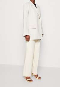 Krémové sako na jedno knoflíku, s širokými rukávy a předními kapsami, ve spojení s krémovými širokými kalhotami a tělovými sandály na vysokém podpatku s pásky.