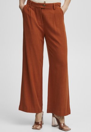 Pantaloni a gamba larga in tessuto arancione bruciato, con chiusura a bottone e pieghe frontali. Abbinati a sandali con tacco trasparente.