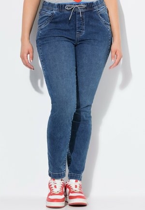 Pantalons jogger en denim, bleu foncé, avec une taille élastique à cordon de serrage, des poches avant et un design de jambe fuselée avec des poignets roulés. Portés avec des baskets rouges et blanches.