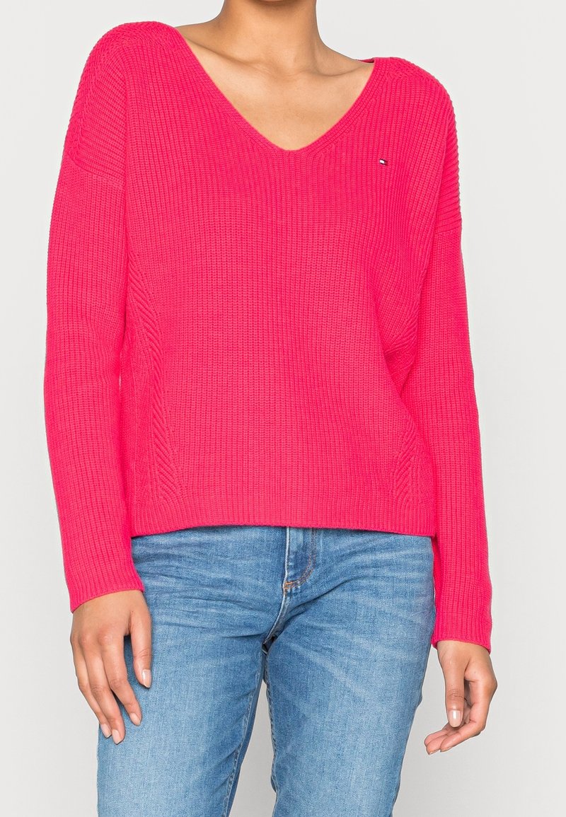 Femme portant un pull en tricot rose vif à col en V et un jean bleu, debout avec les bras détendus contre un fond uni.