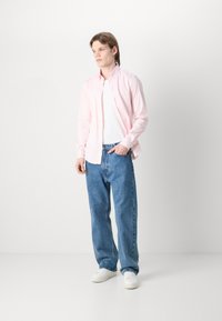 Cămașă roz deschis cu dungi, cu butoni, purtată peste un tricou alb, asortată cu blugi relaxați albaștri și adidași albi. Design simplu cu o croială lejeră.