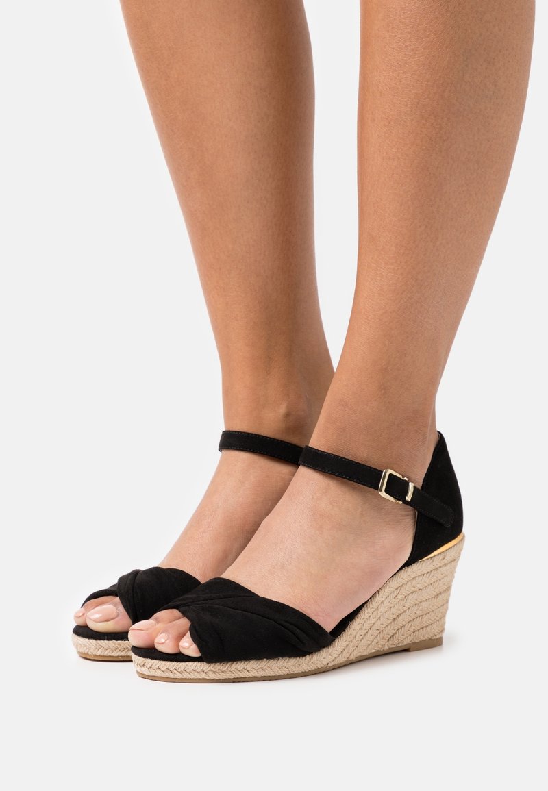 Pieds portant des sandales compensées en daim noir avec des brides à la cheville et des talons enveloppés de jute sur fond blanc.