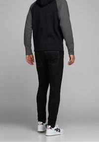 Jeans neri con cuciture arancioni, felpa grigia e blu navy, e sneakers bianche con dettagli neri, viste da dietro.