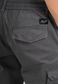 Pantalones cargo grises con cintura elástica, un bolsillo trasero con solapa y una pequeña etiqueta cosida en la cintura. Textura de tela suave.