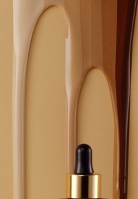 Fonds de teint liquide dans des teintes claires et foncées, avec une texture brillante, goutte à goutte sur une surface beige. Pipette noire avec des accents dorés.