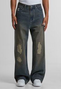 Wide denim traperice u tamnoplavoj boji s istrošenim dijelovima, s pet džepova. Tkanina pokazuje istrošenu teksturu i razderanine na rubovima.