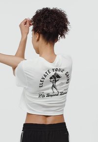 T-shirt blanche raccourcie avec un imprimé graphique noir au dos représentant un haltérophile et le texte "Élévation de vos gains" et "Soulevez au-delà des limites."