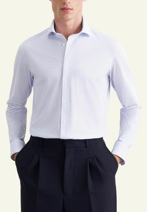 Mężczyzna ubrany w jasnoniebieską teksturowaną koszulę na guziki, włożoną do czarnych plisowanych spodni z rękami w kieszeniach.