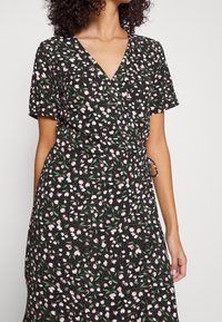 Zwarte jurk met korte mouwen, uitgerust met een wikkeldesign en een bloemenpatroon in roze en groen. De stof heeft een gladde textuur en een vloeiende silhouet.