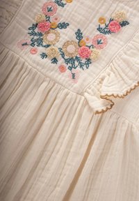 Robe en tissu crème clair ornée de broderies florales colorées en rose, jaune et bleu. Le design comprend des accents volantés et des plis.