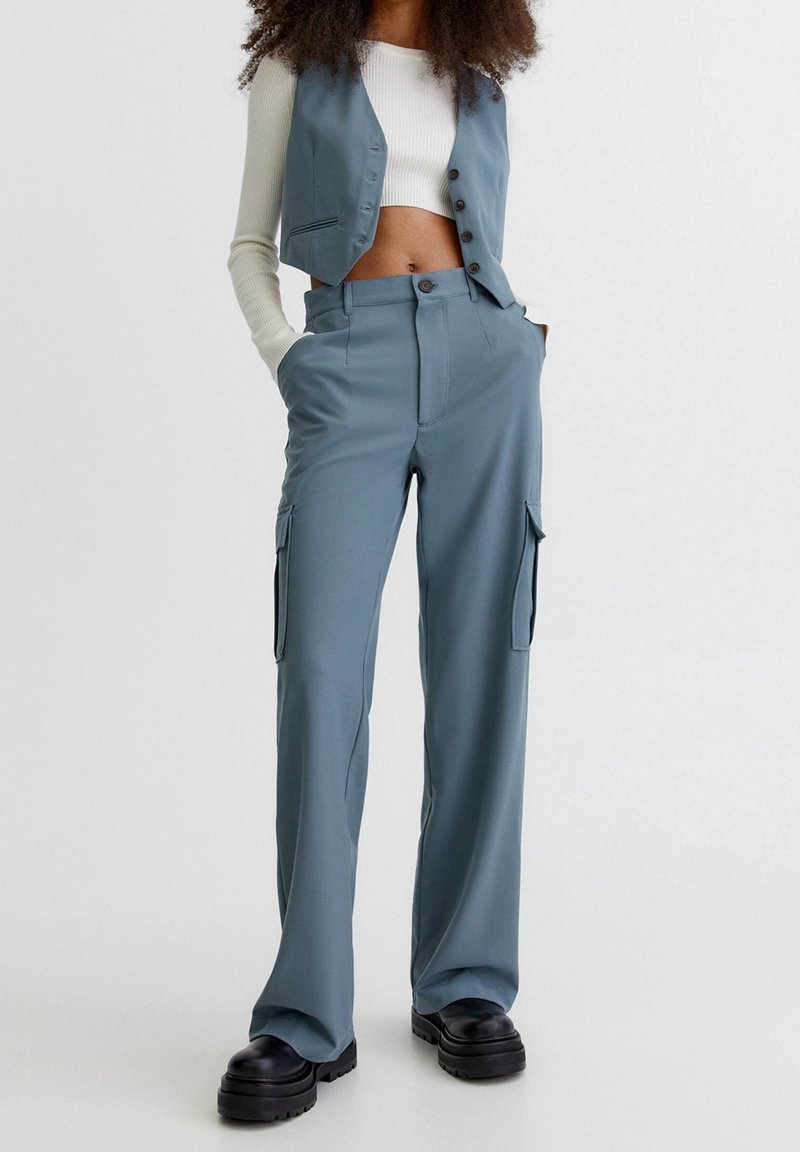 Femme portant un pantalon cargo bleu et un gilet assorti sur un top court à manches longues blanc, debout les mains dans les poches.