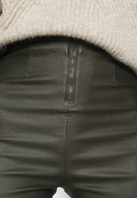 Höga byxor i olivgrön ekologisk läderimitation med en dragkedjedetalj och slät yta, i kombination med en gräddfärgad stickad tröja.