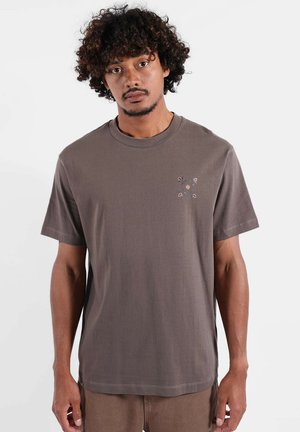 T-shirt en coton marron avec col rond, manches courtes, et un motif floral aux couleurs claires sur la poitrine gauche. Texture lisse.