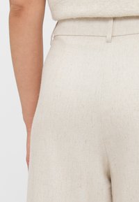 Pantalons beige sur mesure avec une finition texturée, présentant une coupe droite et une taille avec passants de ceinture.