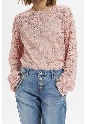 Vrouw die een roze kanten blouse met lange mouwen draagt, ingestopt in een hooggetailleerde blauwe spijkerbroek met vier zichtbare knopen en decoratieve naden.