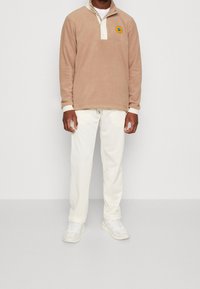 Ljusbrun fleece-pullover med en krämfärgad krage och en cirkulär logotyp. Matchas med vita byxor och vita sportskor.