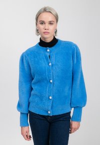 Frau mit kurzen blonden Haaren trägt einen leuchtend blauen, flauschigen Cardigan mit großen Perlenknöpfen über einem schwarzen Rollkragenpullover und dunklen Jeans.