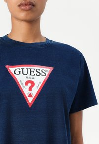 Tamsiai mėlynos spalvos medvilninė marškinėliai su dideliu raudonu ir baltu trikampio logotipu, ant kurio puikuojasi užrašai "GUESS" ir "?". Trumpomis rankovėmis ir apvaliu kaklu.