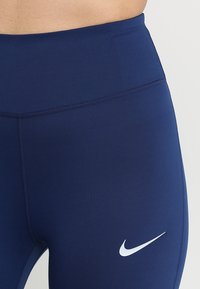 Leggings de sport bleu marine à taille haute, fabriqués en tissu extensible. Présentent un logo Nike blanc et des coutures plates pour plus de confort.