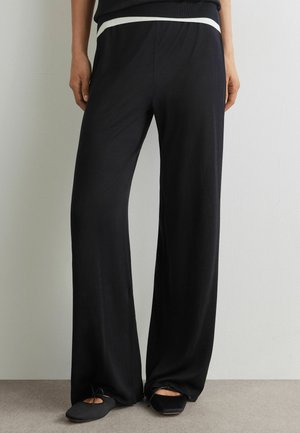 Donna con pantaloni neri larghi con dettaglio in vita bianco e scarpe basse nere, in piedi contro un muro semplice.