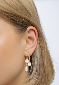 Goldene Ohrringe mit drei hängenden weißen Perlen, die in Form und Größe variieren, befestigt an einem strukturierten runden Haken.